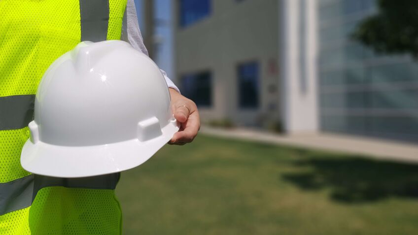 Ein Arbeiter mit Warnweste hält einen weißen Schutzhelm vor sich in der Hand.