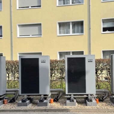 Außenaufnahme von vier grauen Wärmepumpen. Im Hintergrund ist eine Hecke und Hausfassade zu sehen.