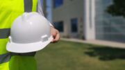 Ein Arbeiter mit Warnweste hält einen weißen Schutzhelm vor sich in der Hand.